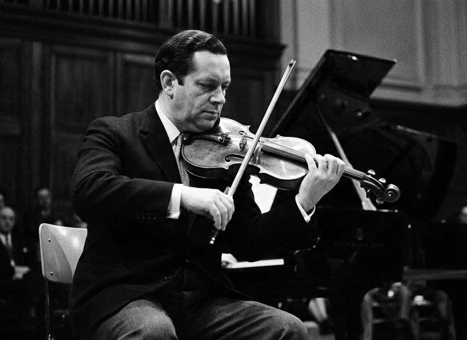 Black-and-white photograph of a seated violinist playing beside a piano